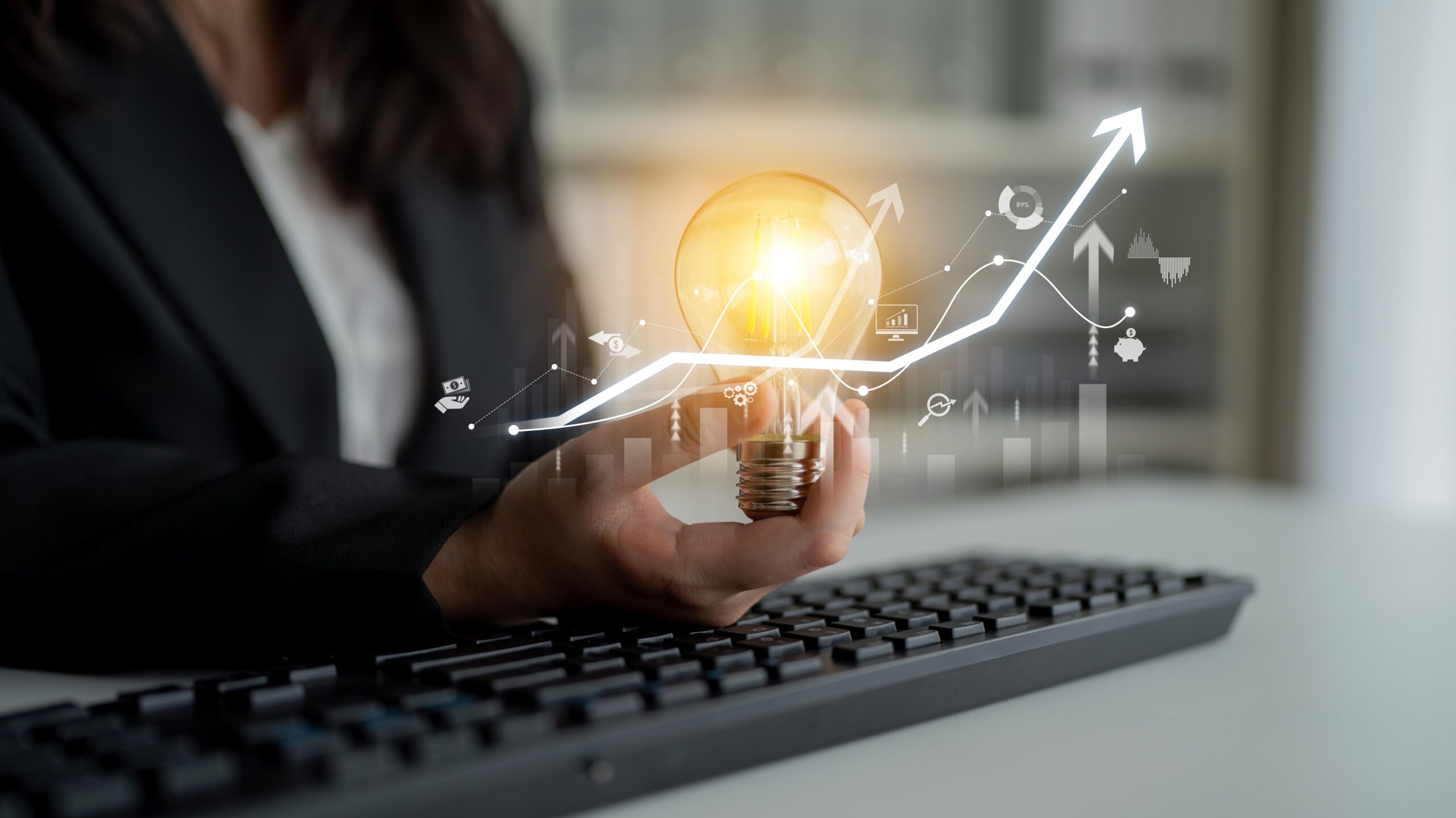 A business professional holds a glowing light bulb while seated at a keyboard, symbolizing innovation and growth, with graphs and icons overlaid.